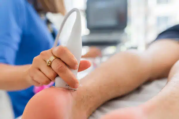 Close up of a surgeon's hand doing an ultrasound of the Achilles tendon on a patient as he lies on an examination table in the office.